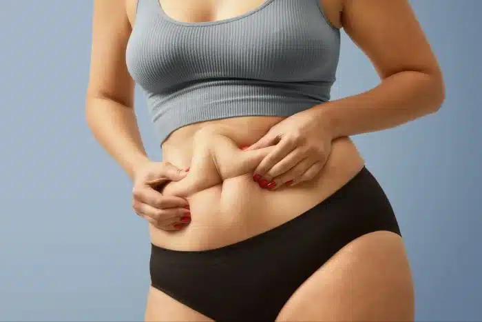 A woman wearing a gray crop top and black underwear pinches the skin on her bare stomach with both hands, showcasing her natural body. The background is a plain light blue.