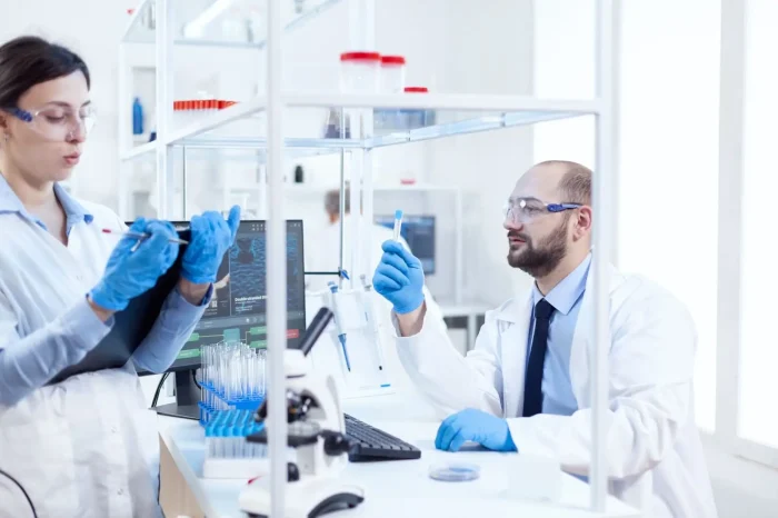 Two scientists in lab coats and safety glasses work in a bright laboratory. One holds a clipboard, the other examines a test tube, with lab equipment and a computer visible on the desk between them.
