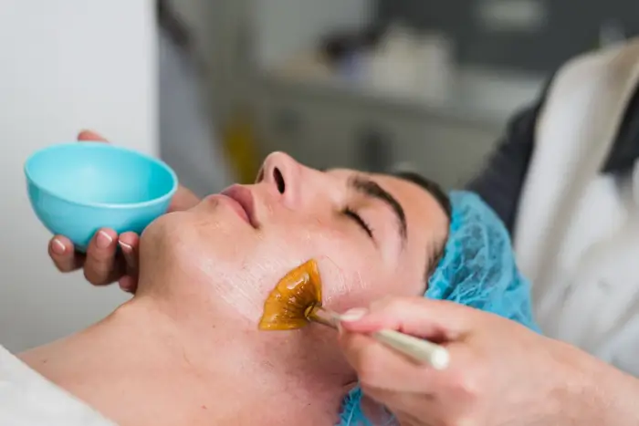 A person lies with eyes closed, wearing a blue hair cap, while another person applies a golden facial mask to their cheek using a brush. A small blue bowl is held nearby.