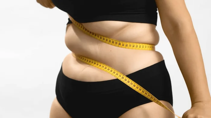 A person wearing a black bra and underwear wraps a yellow measuring tape around their waist, highlighting their midsection against a plain light background.
