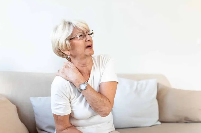 An older woman with short gray hair and glasses sits on a couch, holding her left shoulder and appearing to be in pain or discomfort. She is wearing a white shirt and a watch.